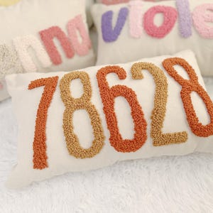 Peut inclure: Un oreiller blanc avec le numéro 78628 brodé en fil orange et marron. Les chiffres sont faits de petits pompons ronds.