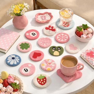 May include: A collection of colorful, handmade crochet coasters in various designs, including a flower, a heart, a strawberry, and a frog. A pink coffee mug sits on a matching coaster. Other items include a vase of flowers, a bowl of strawberries, and a glass of water.