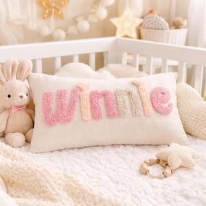 May include: A cream-coloured rectangular cushion embroidered with the name "Winnie" in pink, peach, and beige. A plush bunny and a star-shaped teether are nearby. The cushion is on a white blanket, suggesting a nursery setting.