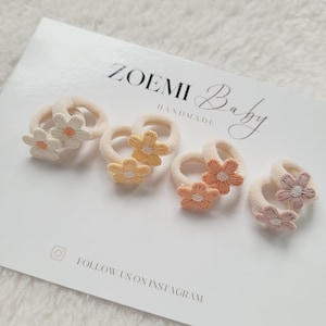 Könnte beinhalten: Satz mit vier Haargummis mit gestickten Blumenakzenten. Die Haargummis sind in den Farben Weiß, Gelb, Orange und Rosa.