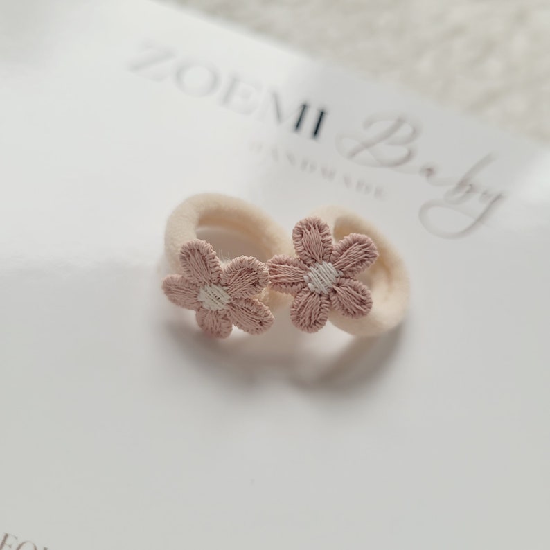 以下が含まれることがあります： 白い中心を持つピンクの花の形をしたヘアゴム2つ。ヘアゴムは柔らかく伸縮性のある素材で作られており、小さな女の子に最適です。