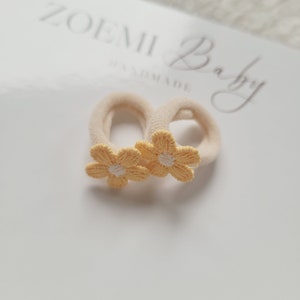 以下が含まれることがあります： クリーム色のヘアゴム2つに、黄色い刺繍の花が施されています。ヘアゴムは、黒い文字で「ZOEMI Baby Handmade」と書かれた白い表面の上に置かれています。