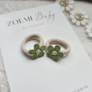 以下が含まれることがあります： 緑の花の装飾が施されたベージュのヘアゴム2つ。ヘアゴムは、"ZOEMI Baby HANDMADE"と印刷された白い表面の上に置かれています。