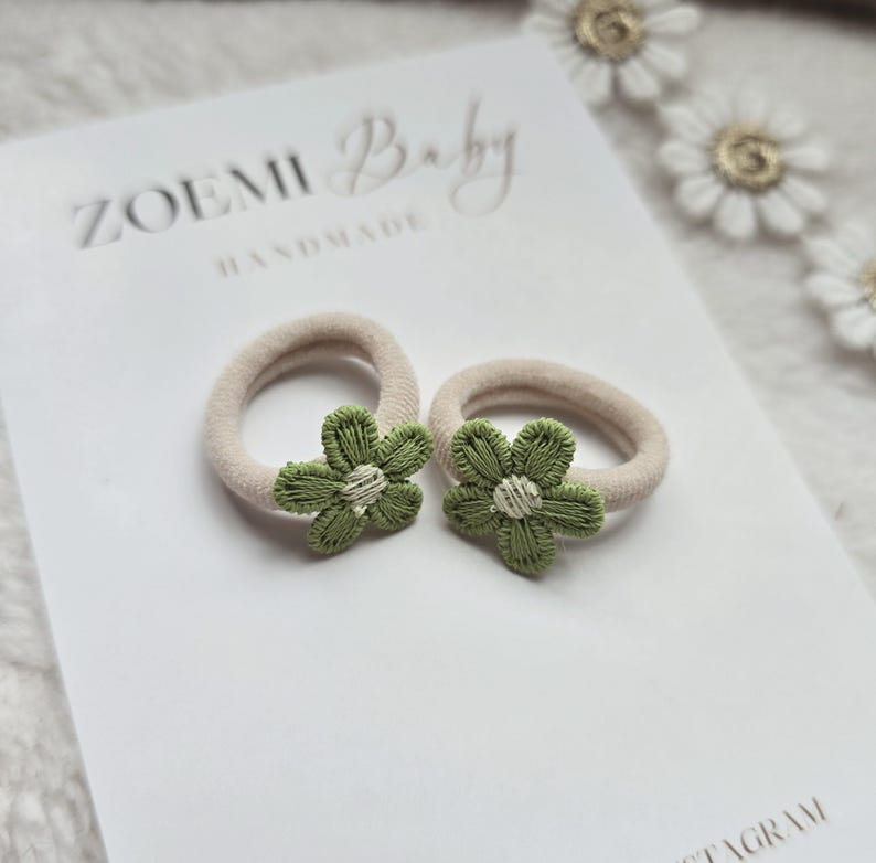 以下が含まれることがあります： 緑の花の装飾が施されたベージュのヘアゴム2つ。ヘアゴムは、"ZOEMI Baby HANDMADE"と印刷された白い表面の上に置かれています。