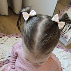 Peut inclure: Un jeune enfant aux cheveux bruns portant un pull rose et deux nœuds blancs dans les cheveux.