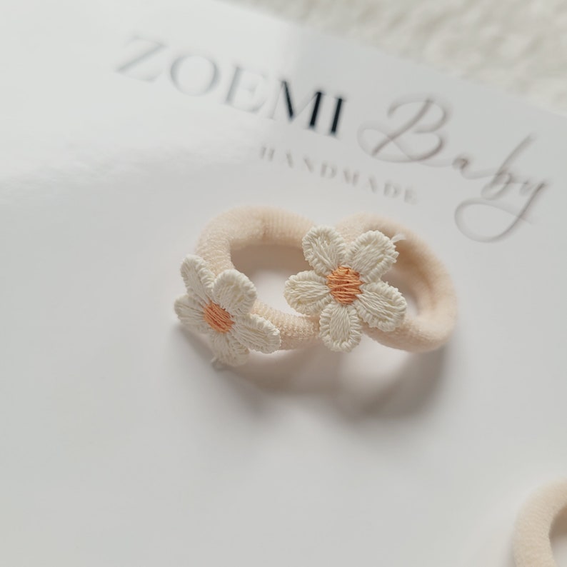 以下が含まれることがあります： 白い刺繍のデイジーの花が付いたベージュのヘアゴム2本。ヘアゴムは、黒い筆記体で「ZOEMI Baby Handmade」と書かれた白い表面の上に置かれています。