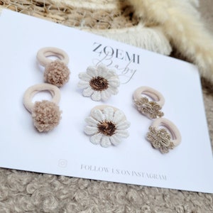 Op de afbeelding: Set van zes haarbandjes met verschillende ontwerpen. Twee haarbandjes hebben een bruine pompon, twee hebben een witte madeliefje met een bruin midden en twee hebben een gouden madeliefje. De haarbandjes liggen op een witte achtergrond met de tekst "ZOEMI Baby" en "FOLLOW US ON INSTAGRAM".