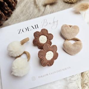 Op de afbeelding: Een verzameling haaraccessoires voor baby's, waaronder twee bruine bloemvormige haarclips met witte centra, twee pluizige witte pompon haarelastiekjes en twee hartvormige haarelastiekjes. De accessoires liggen op een witte kaart met de tekst "ZOEMI Baby".