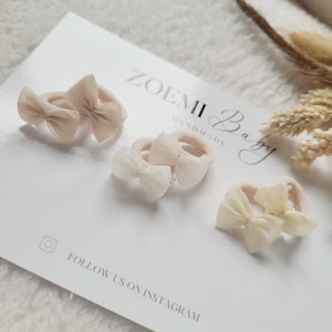 May include: Set of four beige hair ties with bows. The hair ties are handmade and feature the text "Zoemi Baby Handmade".