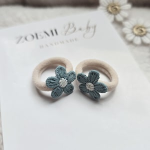 以下が含まれることがあります： 青い花柄の装飾が施されたベージュのヘアゴム2個。花の中心は白く、白い糸で縫われています。