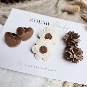 Op de afbeelding: Een collectie haaraccessoires voor baby's. De afbeelding toont twee bruine hartvormige clips, twee witte madeliefvormige clips met bruine centra en twee bruine bloemvormige clips. De merknaam "ZOEMI Baby" is zichtbaar op de kaart.