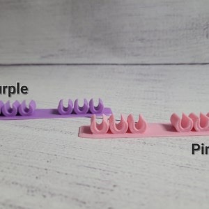K&ouml;nnte beinhalten: Zwei Reihen Kabelorganizer, eine lila und eine rosa. Jede Reihe hat f&uuml;nf Kabelorganizer.