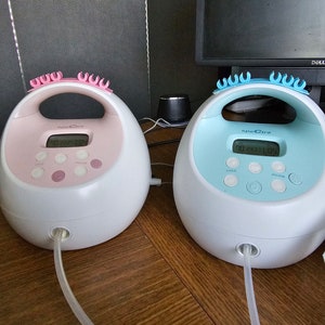 K&ouml;nnte beinhalten: Zwei elektrische Milchpumpen, eine rosa und eine blaue, mit digitalen Displays, die "00 23 31 00" und "00 23 31 05" anzeigen. Beide Pumpen haben einen durchsichtigen Schlauch.