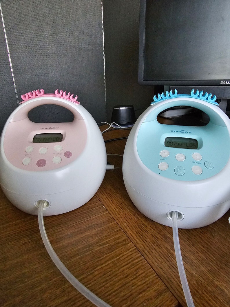 K&ouml;nnte beinhalten: Zwei elektrische Milchpumpen, eine rosa und eine blaue, mit digitalen Displays, die "00 23 31 00" und "00 23 31 05" anzeigen. Beide Pumpen haben einen durchsichtigen Schlauch.