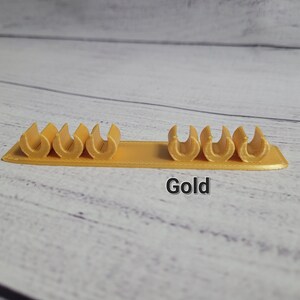 K&ouml;nnte beinhalten: Goldfarbener Kabelorganizer aus Kunststoff mit sechs Schlitzen zum Halten von Kabeln und Dr&auml;hten. Der Organizer befindet sich auf einer hellholzfarbenen Oberfl&auml;che. Gold.