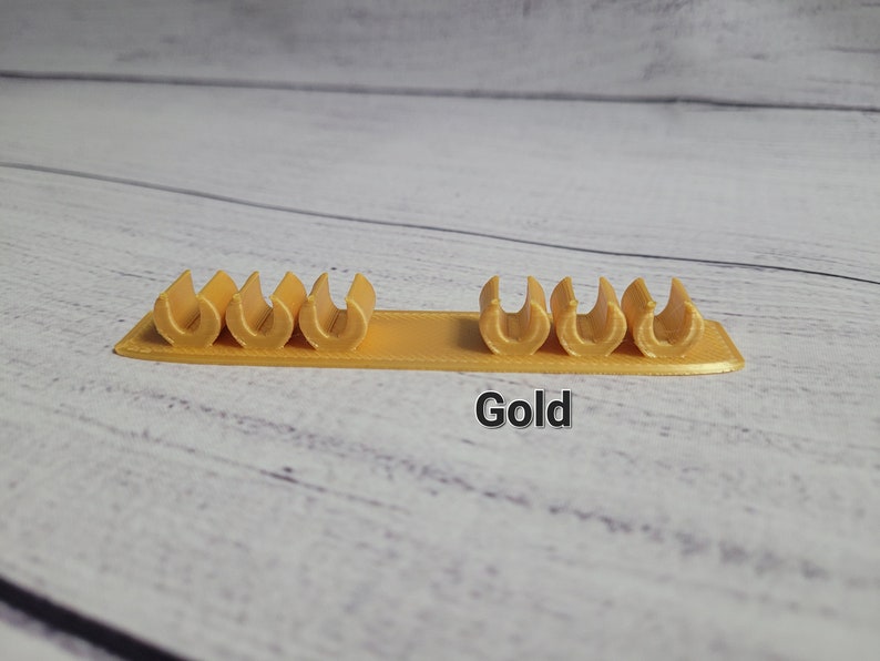 K&ouml;nnte beinhalten: Goldfarbener Kabelorganizer aus Kunststoff mit sechs Schlitzen zum Halten von Kabeln und Dr&auml;hten. Der Organizer befindet sich auf einer hellholzfarbenen Oberfl&auml;che. Gold.