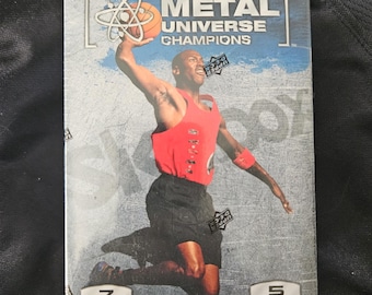 May include: A box of 2021 Skybox Metal Universe Champions basketball trading cards. The box features a basketball player in mid-air, with the text "2021 Skybox Metal Universe Champions" on the front. The box contains 5 packs of cards, with 7 cards per pack.