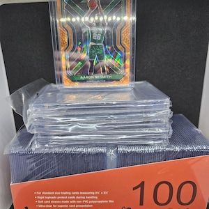 May include: A pack of 100 clear plastic top loaders and card sleeves for standard size trading cards measuring 2 1/2 x 3 1/2 inches. The pack is red with white text that says "100 PACK" and a website address. The image also shows a single trading card in a top loader.