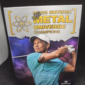 Pode incluir: Uma caixa de cartas colecionáveis da série 2022 Skybox Metal Universe Champions. A caixa apresenta uma foto de um golfista em ação, com o logotipo da Upper Deck e o texto "METAL UNIVERSE CHAMPIONS". A caixa tem um esquema de cores prateado e dourado.