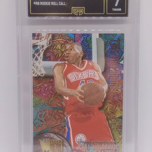 May include: A 1995 Fleer Metal Jerry Stackhouse rookie card graded NM 7. The card features a colorful background with a metallic finish. The card shows Stackhouse in a red and blue Philadelphia 76ers uniform, holding a basketball. The card is graded by GMA.