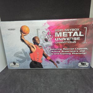 May include: A sealed box of 2021 Skybox Metal Universe Champions trading cards. The box features an image of a basketball player in a red jersey. Text on the box includes "HOBBY", "METAL UNIVERSE CHAMPIONS", and "Featuring Retired Legends".