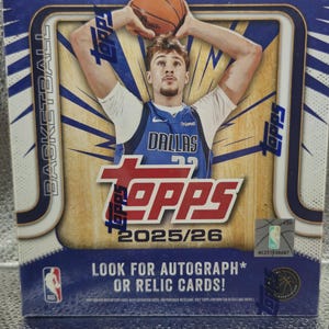 Puede incluir: Una caja de cartas coleccionables de baloncesto Topps, predominantemente azul y dorada, con un jugador con la camiseta de los Dallas Mavericks sosteniendo un balón de baloncesto. La caja presenta el logotipo de Topps y el texto "2025/26" y "Look for Autograph or Relic Cards!"
