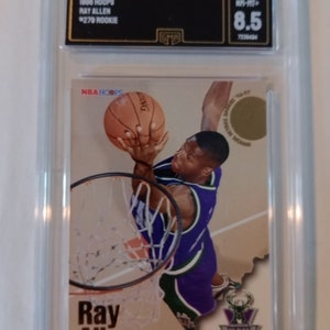 May include: A graded 1998 NBA Hoops trading card featuring Ray Allen of the Milwaukee Bucks. The card is in a protective plastic case with a graded score of 8.5. The card features a photo of Allen in action, with the text "Ray Allen" and "BUCKS" printed on the card.