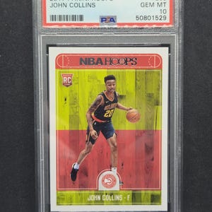 以下が含まれることがあります： 2017 Panini Hoops John Collins ルーキーカード、GEM MT 10 評価、保護ホルダー入り。カードには、アトランタ・ホークスのユニフォームを着てバスケットボールをドリブルするジョン・コリンズの写真が写っています。カードの背景は赤、黄、緑です。