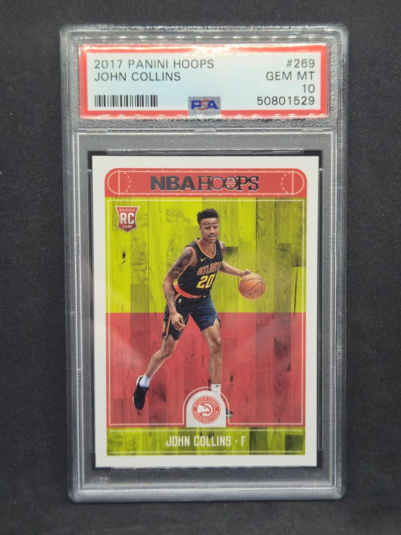 以下が含まれることがあります： 2017 Panini Hoops John Collins ルーキーカード、GEM MT 10 評価、保護ホルダー入り。カードには、アトランタ・ホークスのユニフォームを着てバスケットボールをドリブルするジョン・コリンズの写真が写っています。カードの背景は赤、黄、緑です。