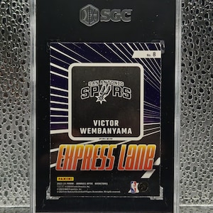 Puede incluir: Una tarjeta de baloncesto Panini Donruss Optic 2023-24 de Victor Wembanyama de los San Antonio Spurs, etiquetada como Express Lane No. 8. La tarjeta est&aacute; encapsulada en una funda protectora SGC negra con frente de pl&aacute;stico transparente y logo plateado.
