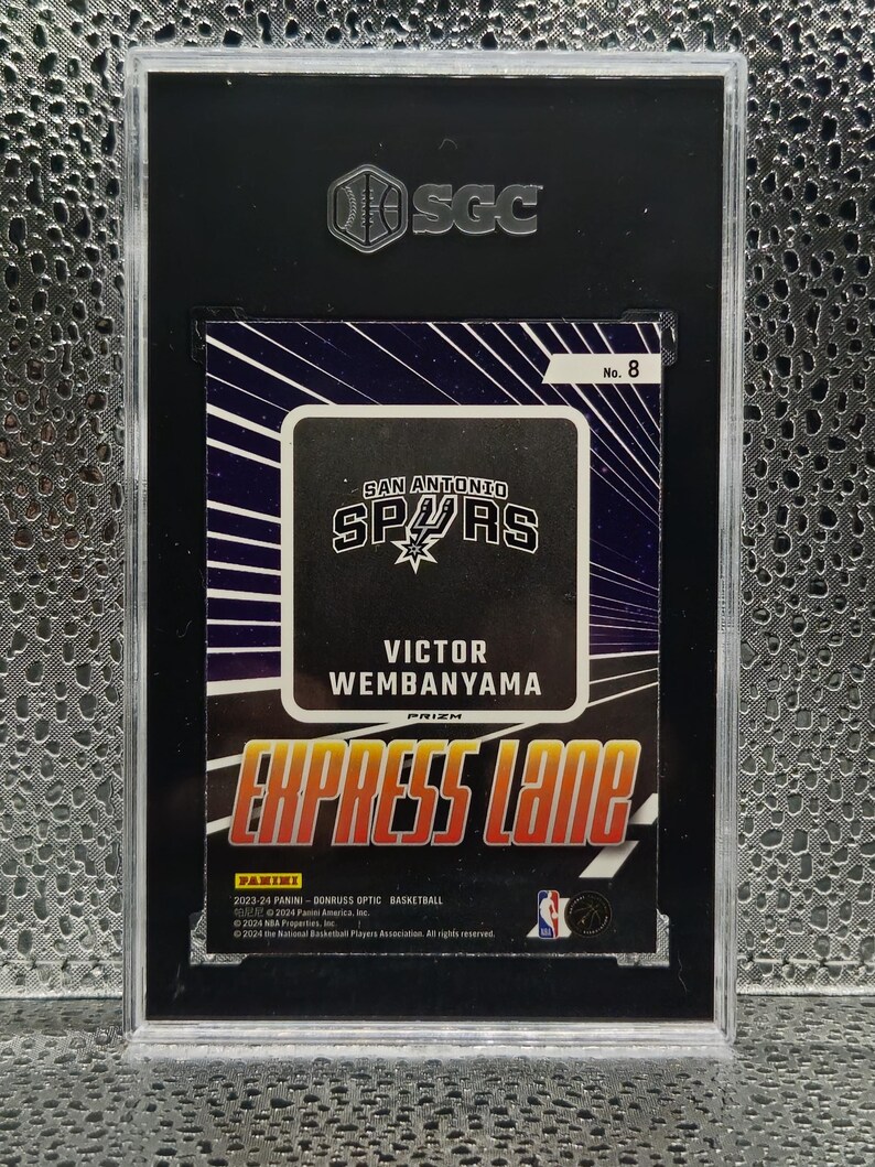 Puede incluir: Una tarjeta de baloncesto Panini Donruss Optic 2023-24 de Victor Wembanyama de los San Antonio Spurs, etiquetada como Express Lane No. 8. La tarjeta est&aacute; encapsulada en una funda protectora SGC negra con frente de pl&aacute;stico transparente y logo plateado.