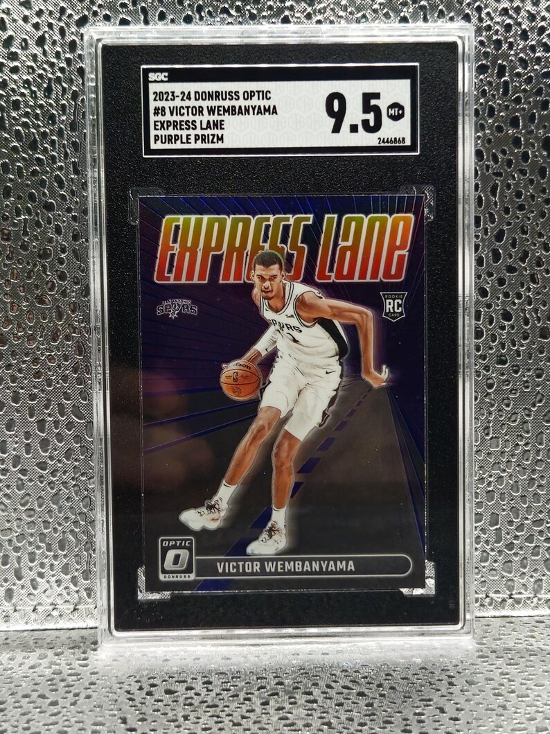 Puede incluir: Una tarjeta de baloncesto 2023-24 Donruss Optic Victor Wembanyama Express Lane Purple Prizm calificada con 9.5 por SGC. La tarjeta muestra al jugador con el uniforme de los San Antonio Spurs driblando un bal&oacute;n sobre un fondo morado con el texto Express Lane.