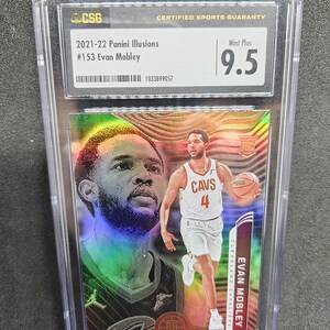 Puede incluir: Una tarjeta de baloncesto Evan Mobley Panini Illusions 2021-22 calificada. La tarjeta est&aacute; encerrada en un soporte de pl&aacute;stico transparente y tiene una calificaci&oacute;n Mint Plus 9.5. La tarjeta presenta una foto de Evan Mobley con su uniforme de los Cavaliers de Cleveland.