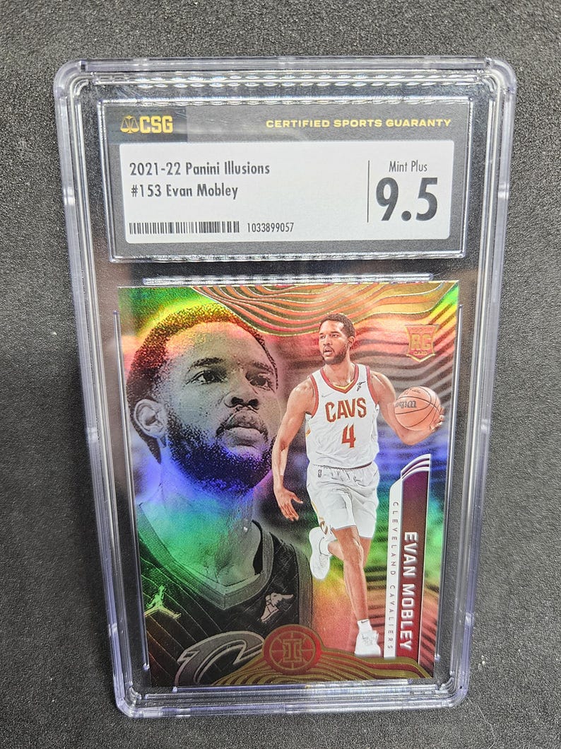 Puede incluir: Una tarjeta de baloncesto Evan Mobley Panini Illusions 2021-22 calificada. La tarjeta est&aacute; encerrada en un soporte de pl&aacute;stico transparente y tiene una calificaci&oacute;n Mint Plus 9.5. La tarjeta presenta una foto de Evan Mobley con su uniforme de los Cavaliers de Cleveland.