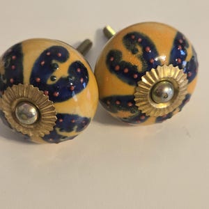 May include: Two round ceramic cabinet knobs featuring a yellow base with dark blue, curved, abstract shapes and small raised orange dots. Each knob has a central gold-toned metal rosette with a silver-colored screw head and a metal mounting bolt.
