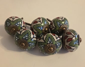 Może przedstawiać: Zestaw pięciu okrągłych ceramicznych gałek do szuflad z ręcznie malowanym wzorem botanicznym przedstawiającym zielone liście, czerwone jagody i niebieskie akcenty na białym tle. Każda gałka ma centralną mosiężną nasadkę i metalową śrubę.
