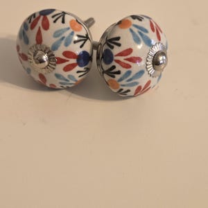 Puede incluir: Dos pomos redondos de cerámica para armarios con base blanca y un patrón floral repetitivo en rojo, azul claro, naranja y azul oscuro. Cada pomo tiene una tapa central de metal plateado y un tornillo metálico roscado en la parte posterior.