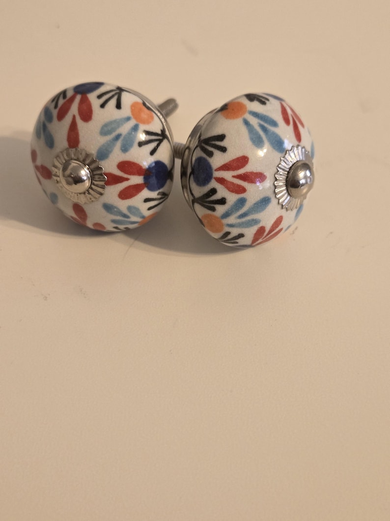 Może przedstawiać: Dwie okrągłe ceramiczne gałki do mebli z białym tłem i powtarzającym się kwiatowym wzorem w kolorach czerwonym, jasnoniebieskim, pomarańczowym i ciemnoniebieskim. Każda gałka ma srebrną metalową nasadkę i gwintowaną śrubę z tyłu.