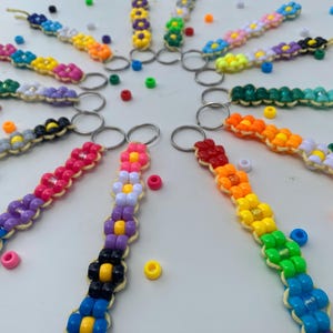 May include: A collection of colorful beaded keychains arranged in a circle. Each keychain features a flower design with various colors, including blue, pink, yellow, and green. The keychains are on a white surface, with scattered beads.
