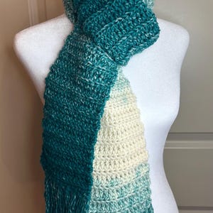 May include: A crocheted scarf with a teal, white, and light blue ombre pattern. The scarf has long fringe on the ends.