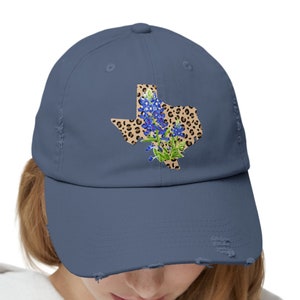 May include: A distressed denim blue baseball cap with a distressed brim. The cap features a  leopard print outline of the state of Texas with bluebonnet flowers.