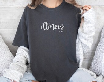 May include: A dark gray t-shirt with the word "illinois" written in white cursive lettering and the text "est. 1818" underneath.