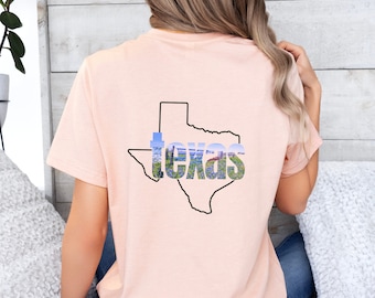 May include: A light peach-colored t-shirt featuring a black outline of the state of Texas. The state outline is filled with a scenic image and the word "Texas" in a stylized font.