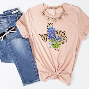 May include: A peach-colored t-shirt with a graphic of the state of Texas in a leopard print design with bluebonnet flowers.