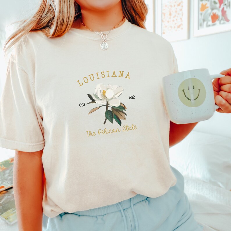 May include: Cream-colored t-shirt featuring a magnolia flower graphic with the text "LOUISIANA" and "The Pelican State." A white mug with a smiley face is held in the hand. The shirt has a relaxed fit.
