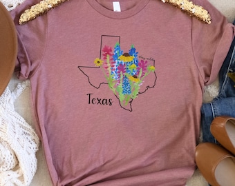 May include: A mauve t-shirt with a graphic of the state of Texas outlined in black with colorful wildflowers inside. The text "stay wild" is written above the flowers and "Texas" is written below the outline of the state.