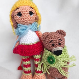 Puede incluir: Una muñeca de crochet con un sombrero amarillo, una falda roja y pantalones a rayas verdes y blancas está de pie junto a un oso de crochet marrón que lleva una bufanda verde con un botón verde.