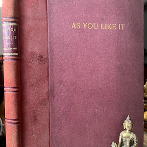May include: A burgundy hardback book with gold lettering on the spine and front cover. The title of the book is "As You Like It" by William Shakespeare. A small gold statue of Buddha is in the foreground.