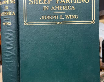 Puede incluir: Un libro de tapa dura verde titulado "Sheep Farming in America" de Joseph E. Wing. El libro tiene letras doradas y un borde dorado alrededor del título. El libro está sobre una superficie de madera.