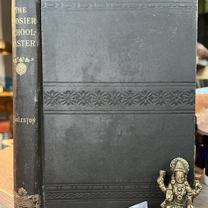 May include: A black hardcover book with gold lettering on the spine. The title is "The Hoosier School-Master" by Eggleston. The book is sitting on a wooden surface with a gold statue of a goddess and a clear crystal.